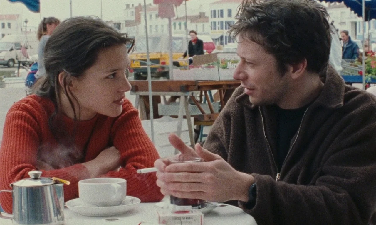 Late August, Early September (1998) movie still: medium — An outdoor table at a street market features a woman and man talking. They sit at a round…
