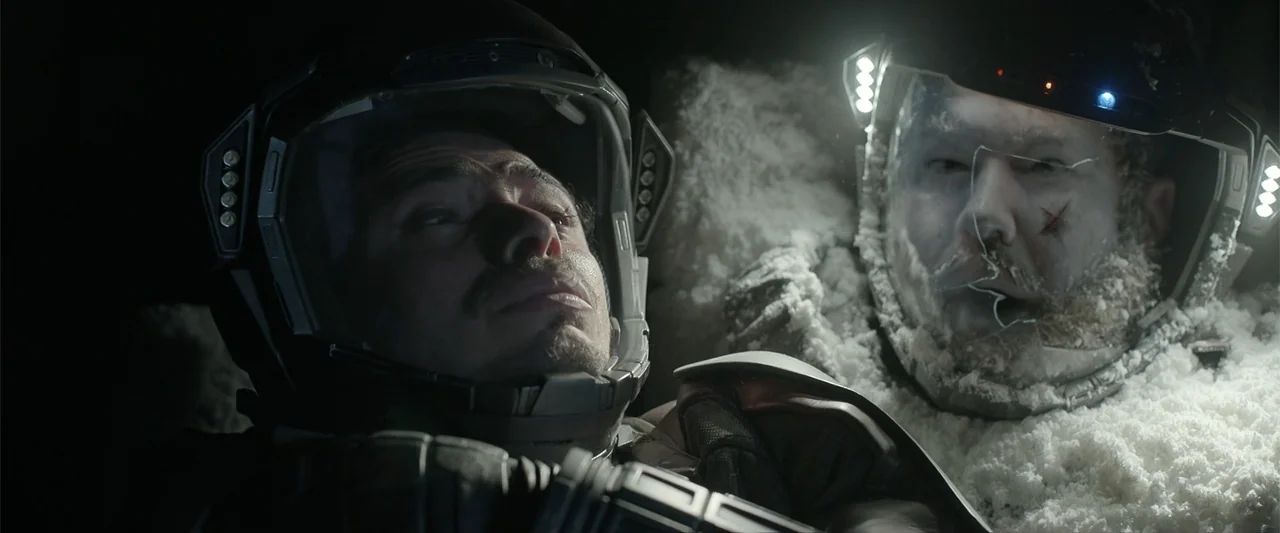 DISTANT (2026) movie still: medium — Two men in bulky futuristic spacesuits lie in a cramped dark enclosure. Their helmets are covered in frozen ice…