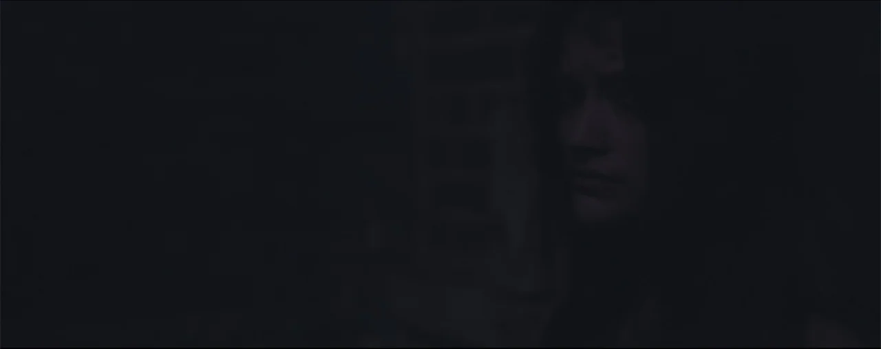 El Último Juego (2025) movie still: closeup — A very dark interior space with a person partially obscured in deep shadow. A woman stares from the…