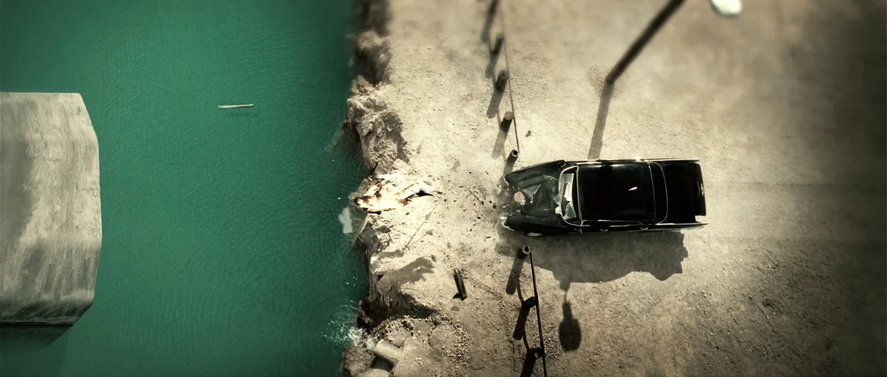 Daybreakers (2009) movie still: establishing — A dark vintage car sits precariously on a dusty cliff edge above green water. The vehicle appears…