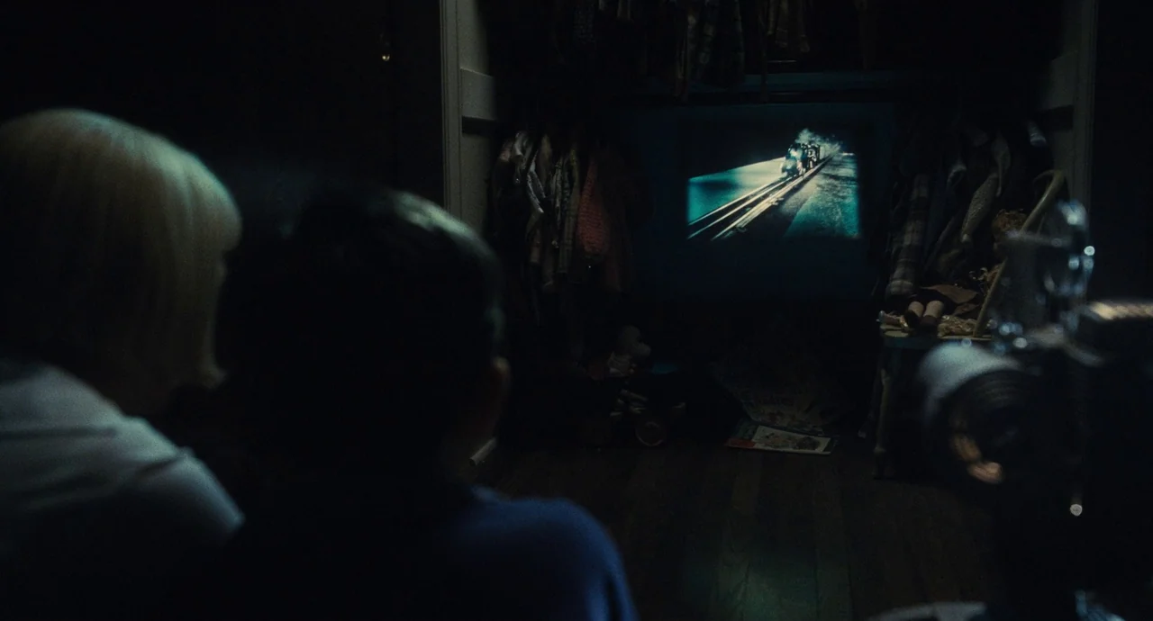 The Fabelmans (2022) movie still: medium — A dark closet contains hanging clothes and a projected film of a train. Two individuals sit in the…