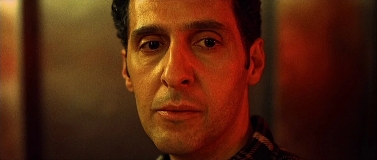 Fear X (2003) movie still: closeup — The interior of an elevator illuminated by a single source of vibrant red light. A man looks directly toward…