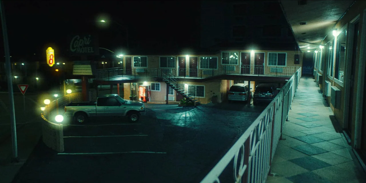 Spermworld (2024) movie still: wide — A two-story roadside motel at night is lit by overhead lamps and neon signs. Two cars are parked in the lot…