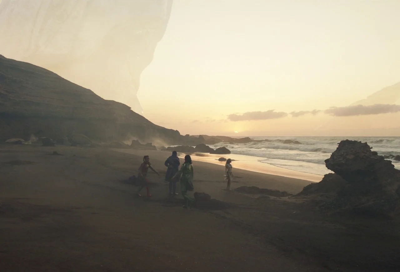 Eternals (2021) movie still: wide — A flat sandy beach at golden hour with rocky cliffs on the left. Four figures in patterned suits stand together…