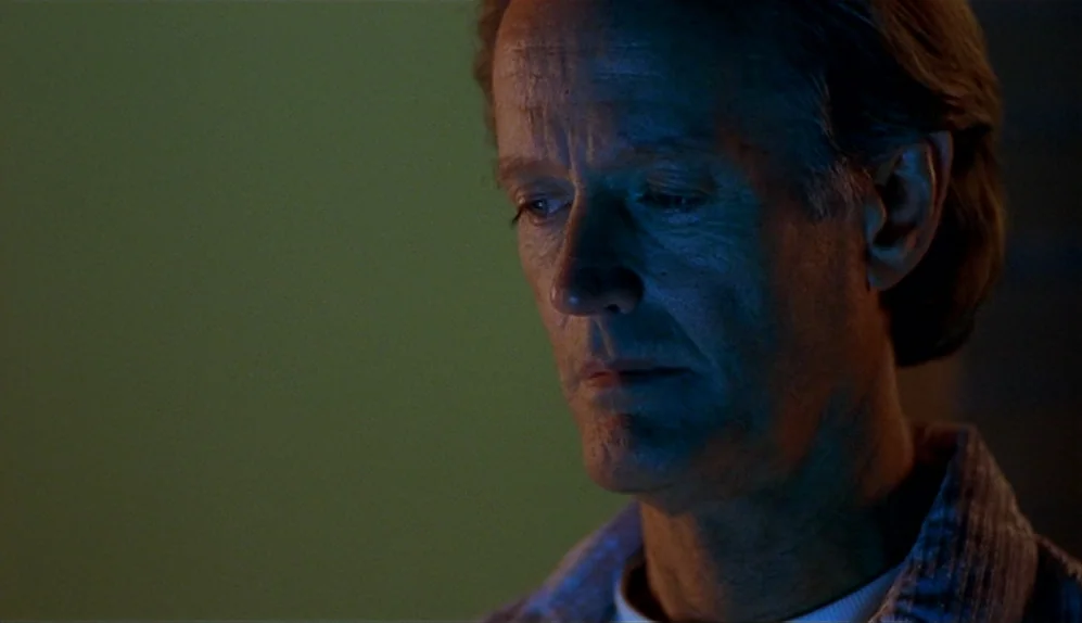 The Limey (1999) movie still: closeup — A man stands in a dimly lit room with muted wall tones. The man gazes downward with a neutral expression.