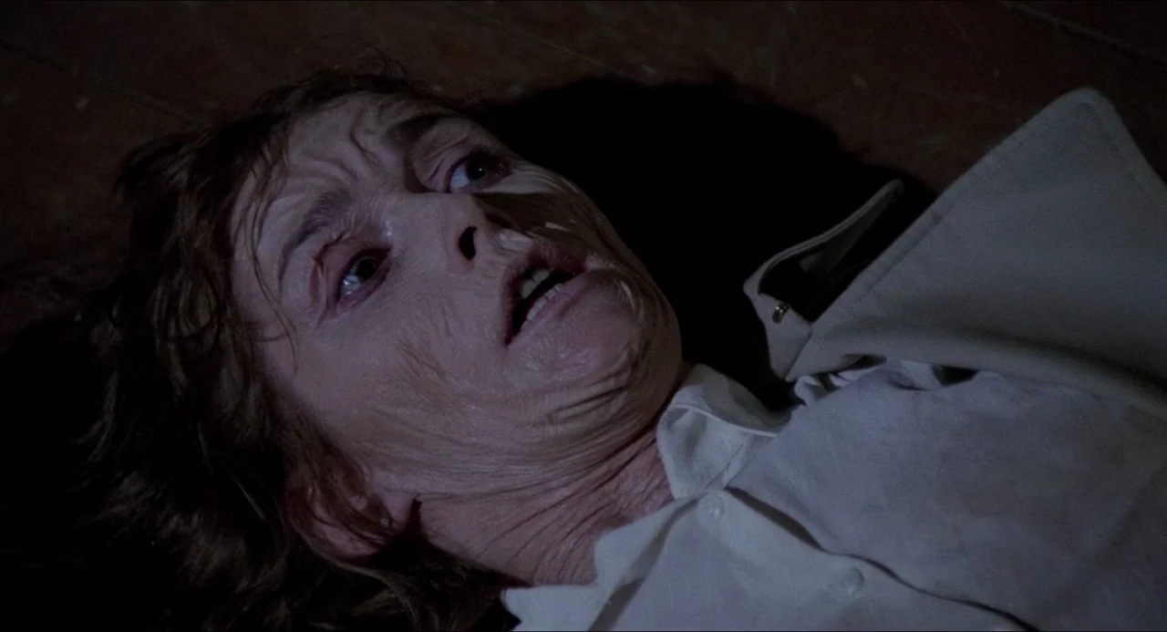 The Amityville Horror (1979) movie still: closeup — An elderly woman lies recumbent on a dark wooden floor in a dimly lit area. She stares upward…