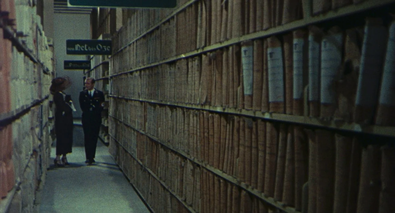 The Damned (1969) movie still: wide — A long, narrow aisle is lined with high industrial shelves packed with thick paper folders. A man in a uniform…