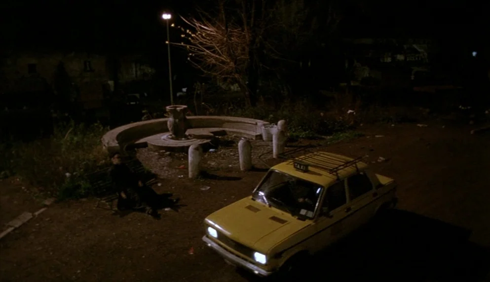 Night on Earth (1991) movie still: wide — An empty urban square at night features a curved concrete fountain and a lamp. A lone man sits on a park…