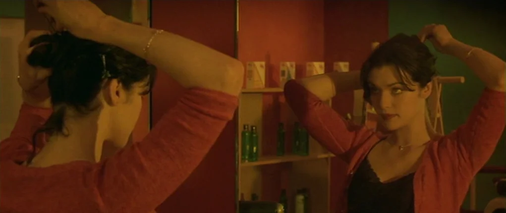 I Want You (1998) movie still: medium — A dimly lit room features a woman looking into a rectangular mirror. She uses both hands to tuck and adjust…
