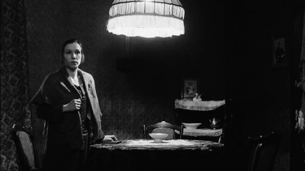 The Role (2013) movie still: medium — A dim room features patterned wallpaper and antique furniture illuminated by a singular pendant lamp. A woman…