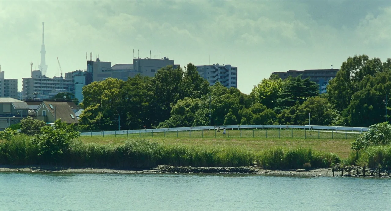 Shoplifters (2018) movie still: wide — A river flows past a grassy bank with a white guardrail and dense trees. The urban skyline includes…