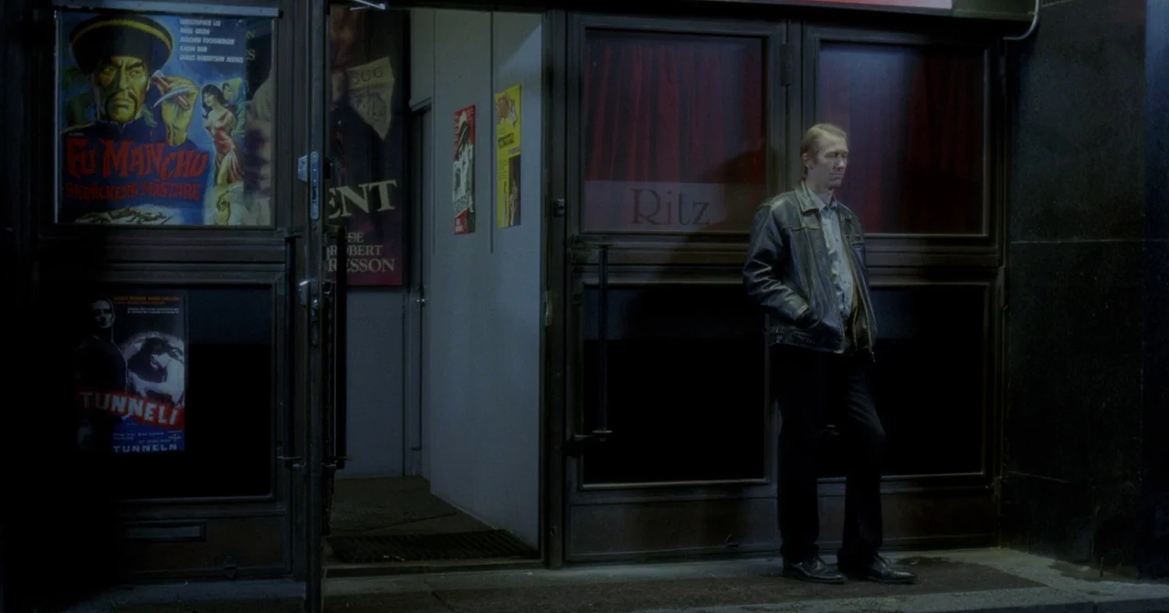 Fallen Leaves (2023) movie still: wide — The exterior of a theater entrance at night features movie posters on glass panels. A man stands motionless…