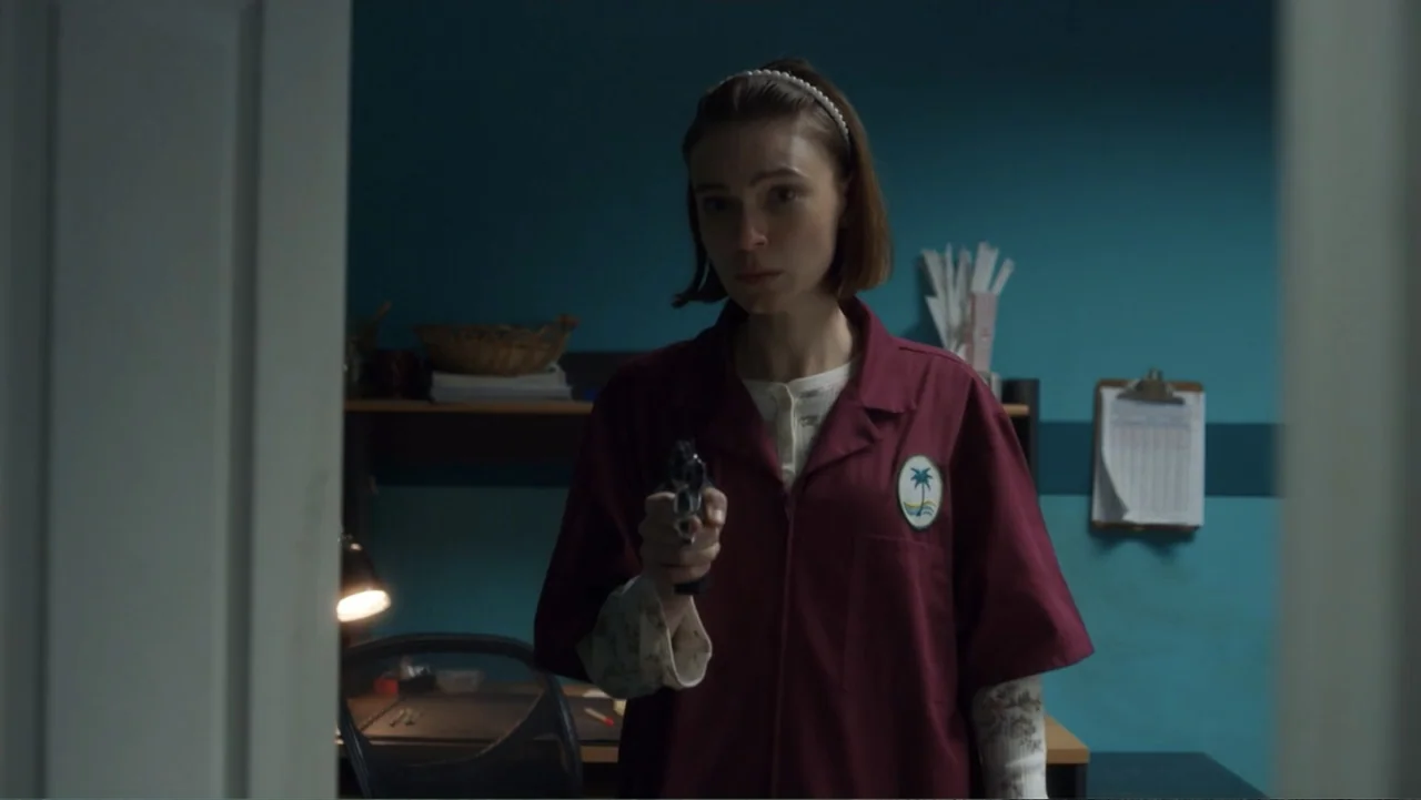 Burn (2019) movie still: medium — A dimly lit room with teal walls features a desk, a wicker basket, and a clipboard. A woman in a work uniform…