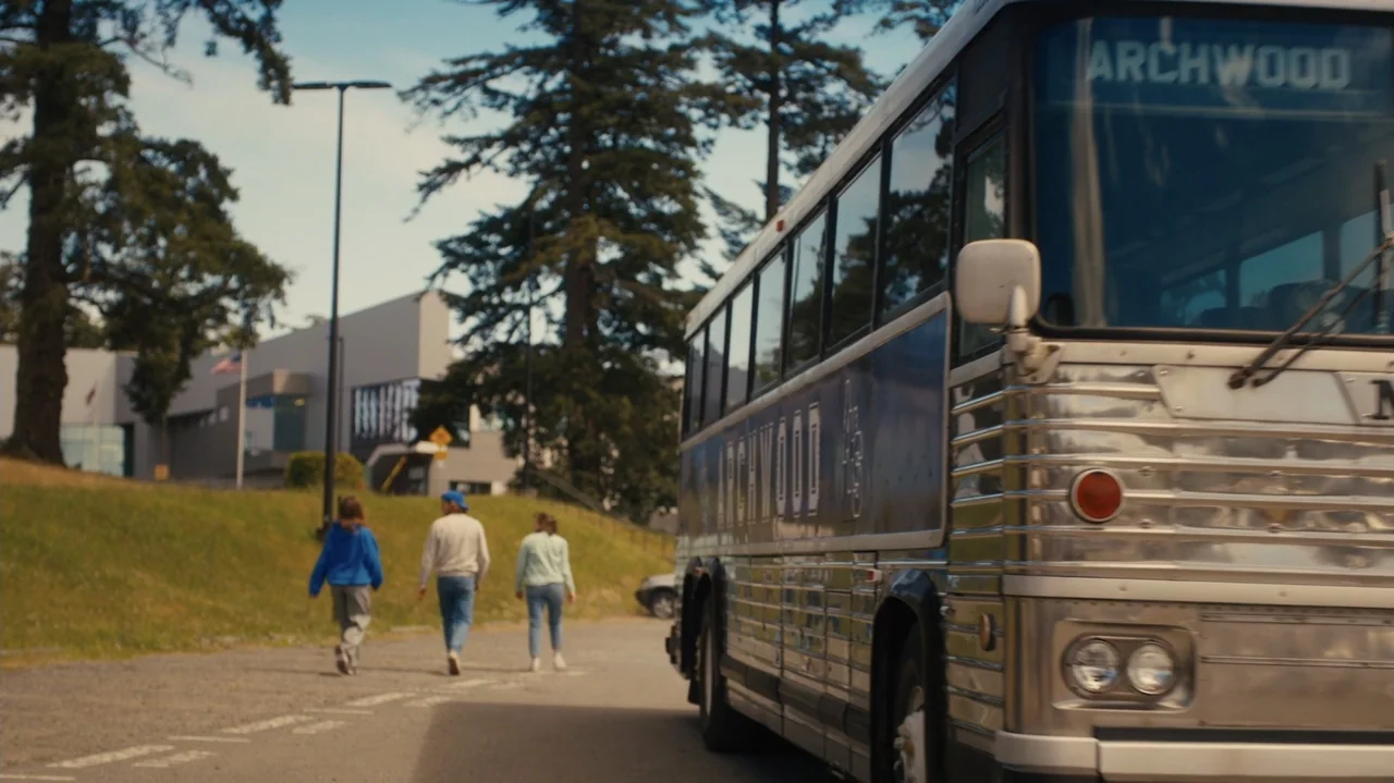Sidelined: The QB and Me (2025) movie still: wide — An asphalt road runs alongside a large vintage bus with a silver metal body. Three people walk…