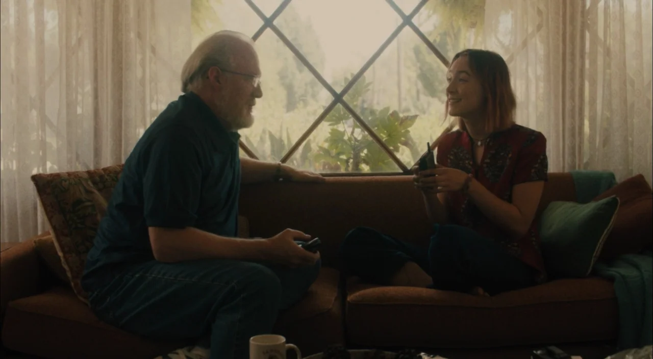 Lady Bird (2017) movie still: medium — A sunny interior space featuring a brown sofa set against a diamond-paned window. An older man and a young…