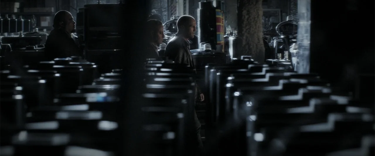 Wild Card (2015) movie still: medium — A dark, dimly lit industrial space is filled with rows of black metal containers. Three men walk between the…
