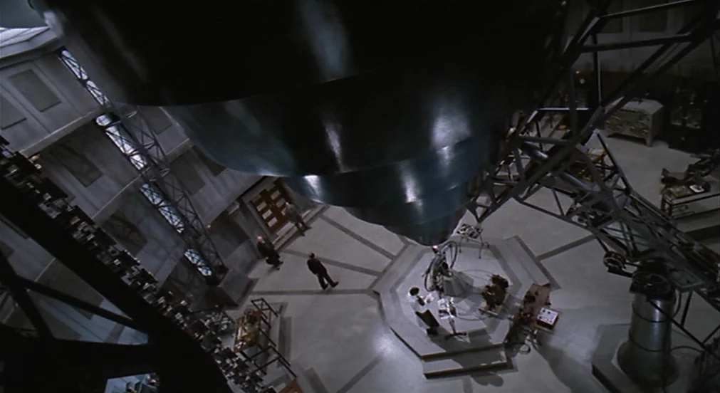Kafka (1991) movie still: wide — A large industrial laboratory features a prominent conical structure suspended from the ceiling. Two men in suits…