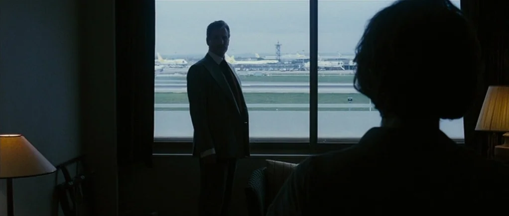 The Walker (2007) movie still: medium — A dimly lit hotel room looks out onto an active airport tarmac with airplanes. A man in a suit stands by the…
