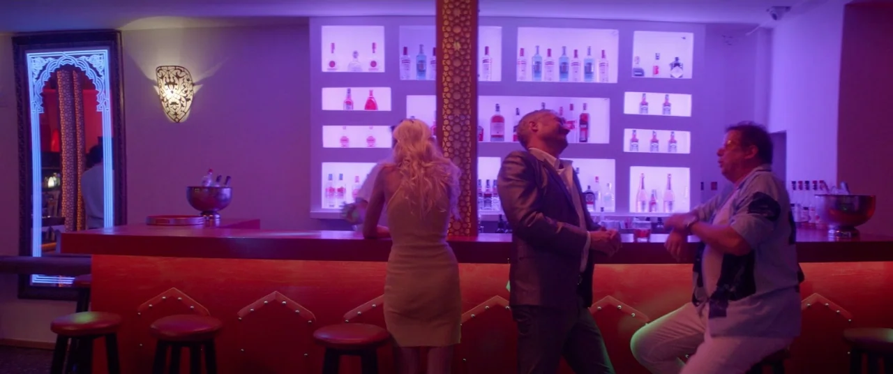 Holiday movie still: wide — A modern bar interior features red counter seating and shelves lined with glass bottles. Three people interact while…