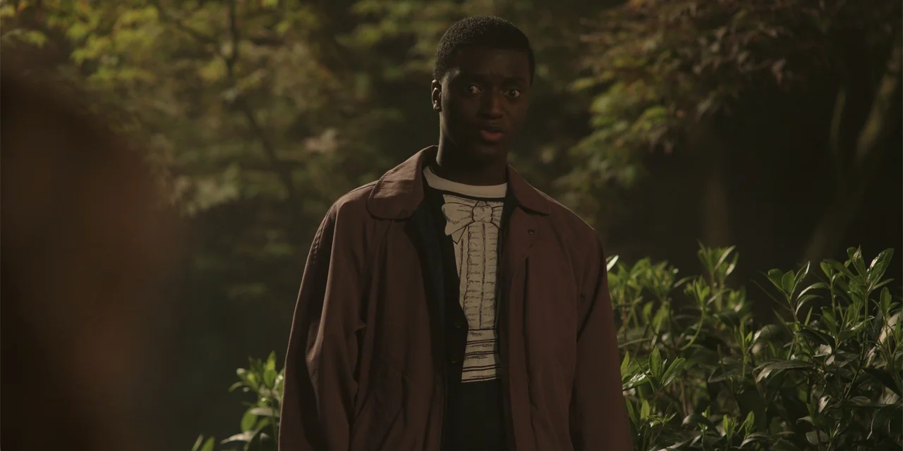 Totally Killer (2023) movie still: medium — A young man stands in a dark, outdoor setting surrounded by dense foliage at night. The man gazes…
