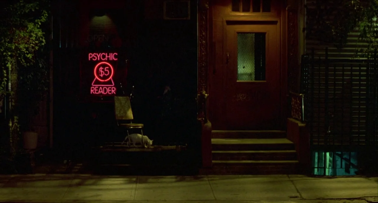 In The Cut (2003) movie still: wide — A dark urban exterior features a building entrance with steps and a metal gate. A bright pink neon psychic…