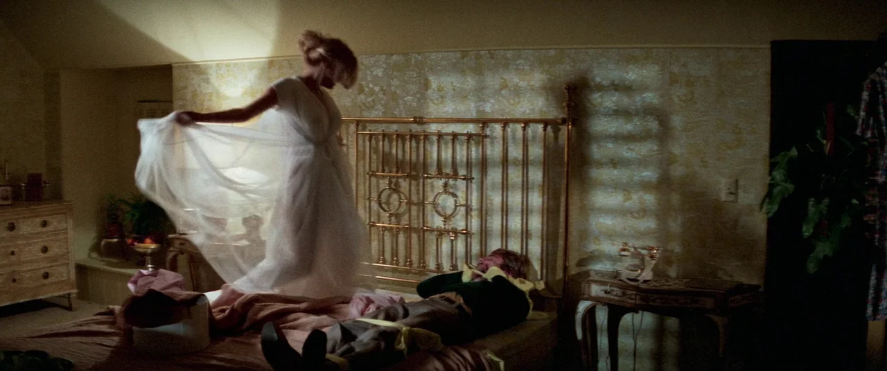 Death Game (1977) movie still: medium — A vintage bedroom features patterned floral wallpaper and a prominent brass-framed headboard. A woman stands…