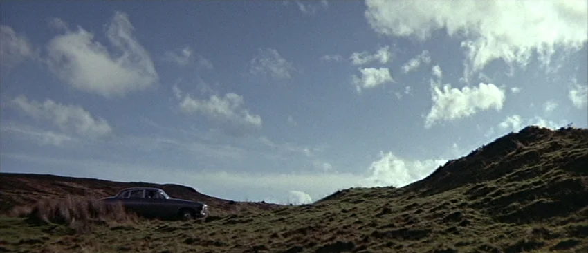 Images (1972) movie still: wide — A rural hilly landscape with patches of dry grass under a partially cloudy sky. A dark sedan is parked on the…