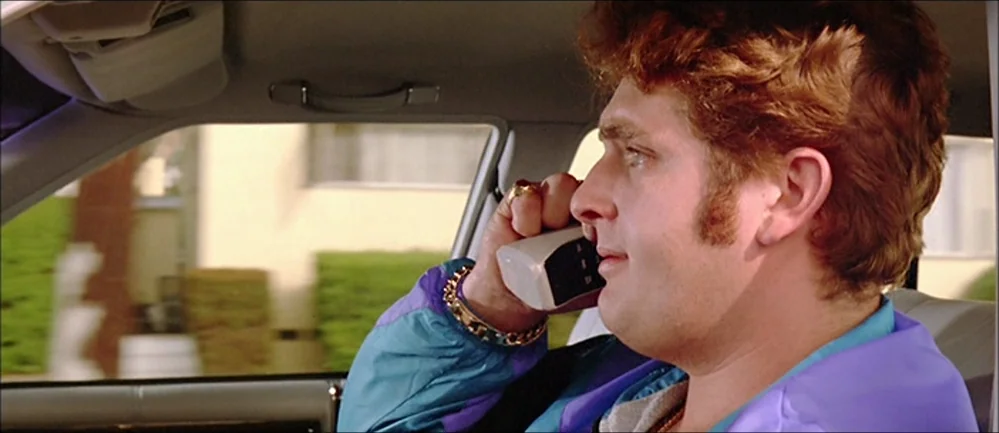 Reservoir Dogs (1992) movie still: medium — The interior of a moving 1980s sedan with beige upholstery. A man with curly red hair talks on a large…