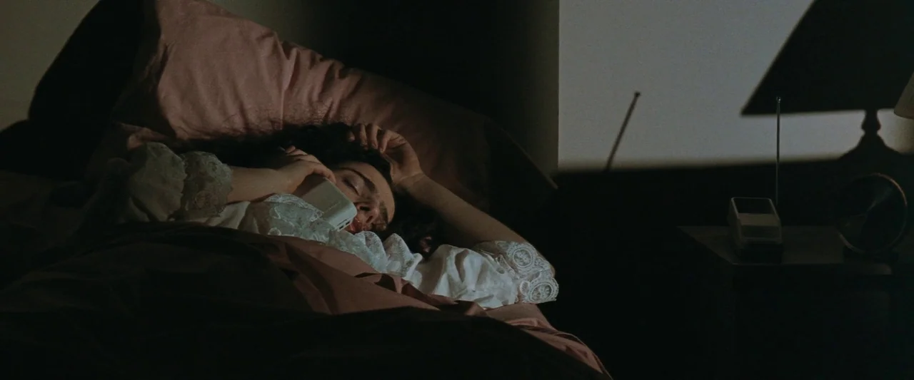 The Adjuster (1991) movie still: medium — A person lies in a dark room resting on pink pillows in a bed. She holds a white telephone handset to her…