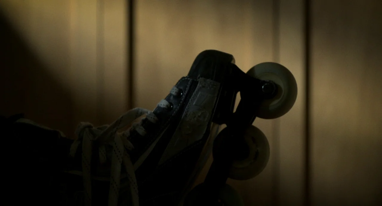 Happer’s Comet (2022) movie still: closeup — A close-up view of an inline skate positioned against a vertical wood-paneled wall. The skate is…