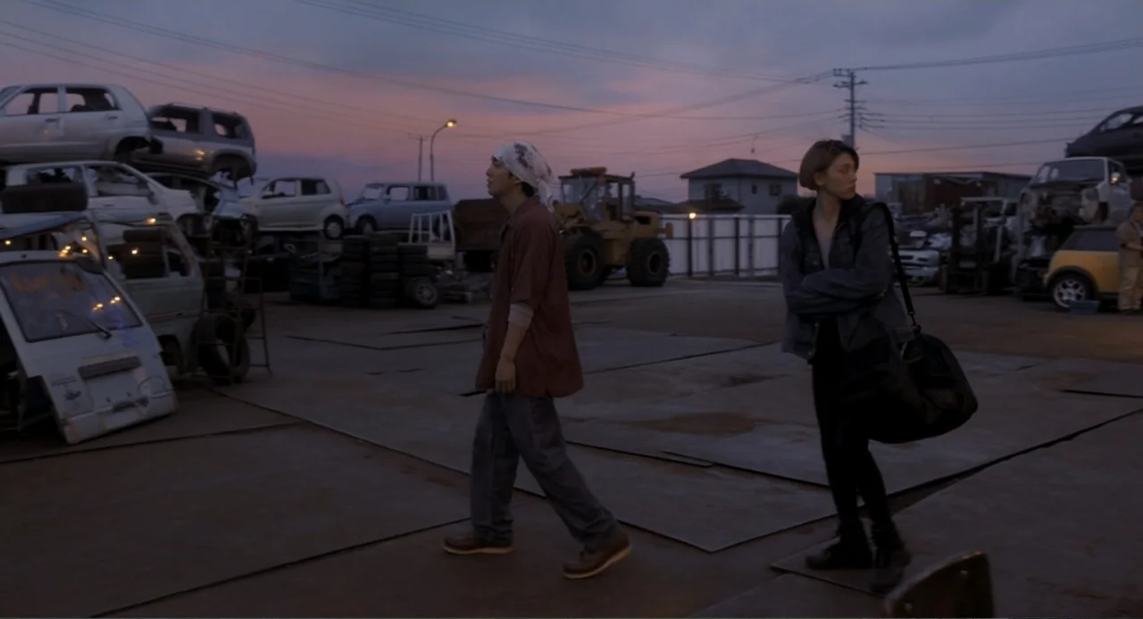 Knuckle Girl (2023) movie still: fullbody — A sprawling outdoor vehicle scrapyard under a purple and orange sunset sky. A young man walks past a…