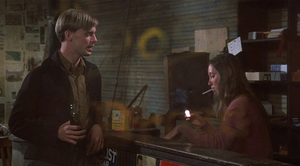 Thieves Like Us (1974) movie still: medium — A cluttered 1970s general store interior has wooden shelves and corrugated metal wall panels. A man…
