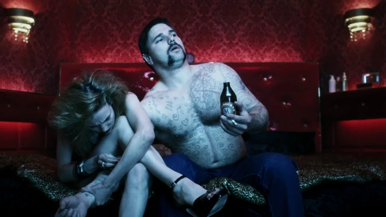 Chopper (2000) movie still: medium — A dimly lit room features deep red walls covered in damask-style patterned wallpaper. A man with tattoos sits…