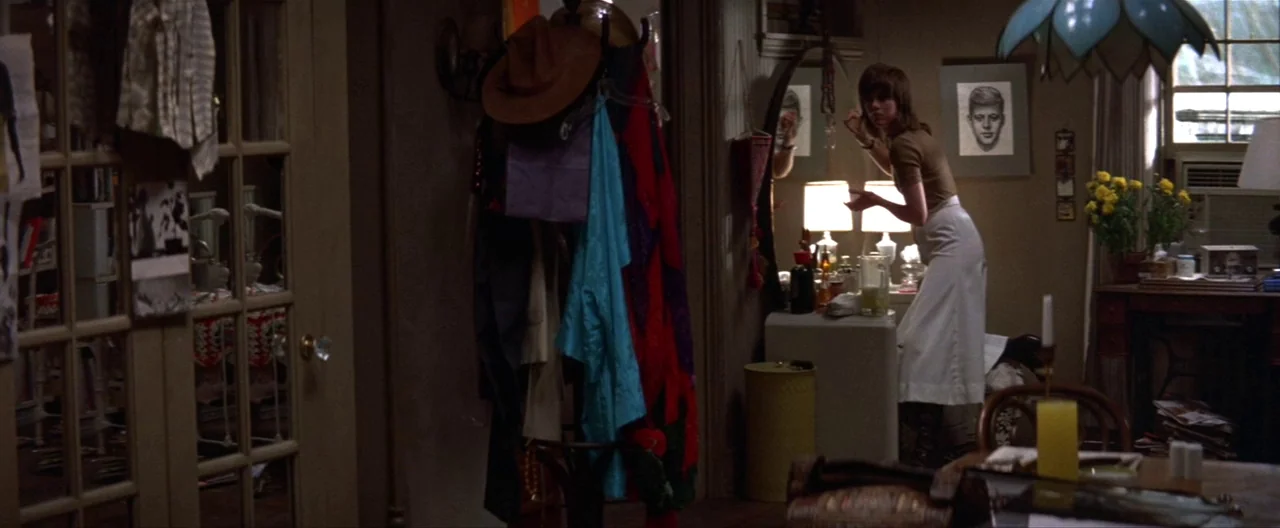 Klute (1971) movie still: wide — A cluttered apartment interior with clothes hanging on a rack near a window. A woman stands in front of a mirror…