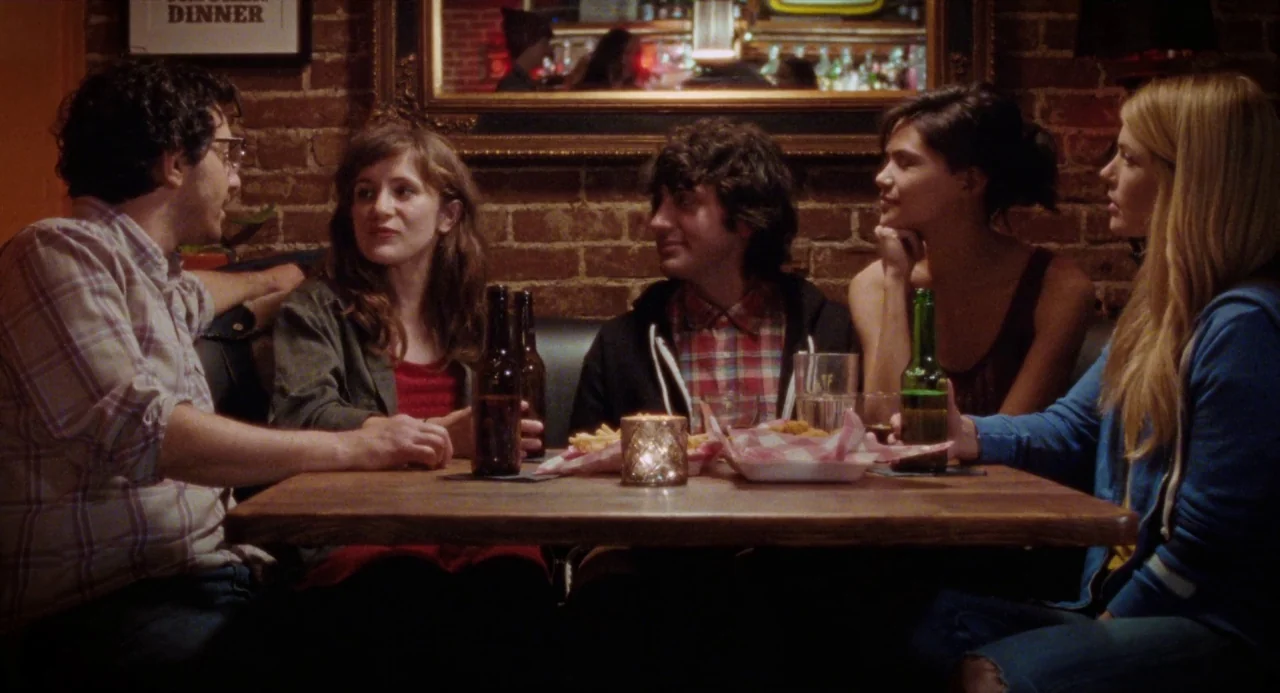 Mr. Roosevelt (2017) movie still: medium — Four people sit inside a brick-walled bar booth around a wooden table. They are engaged in conversation…