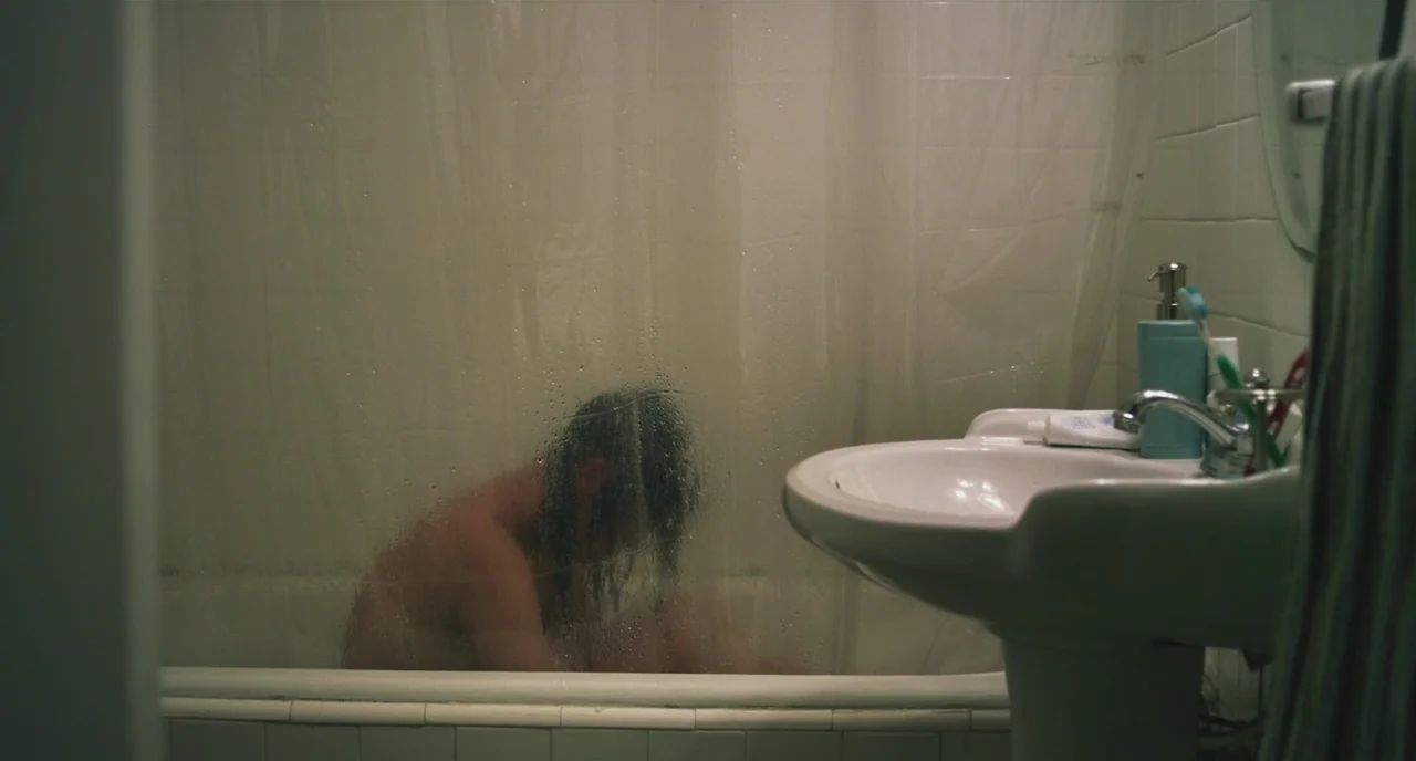 Short Term 12 (2013) movie still: medium — A dimly lit room features a pedestal sink beside a shower with a curtain. A figure sits huddled within…