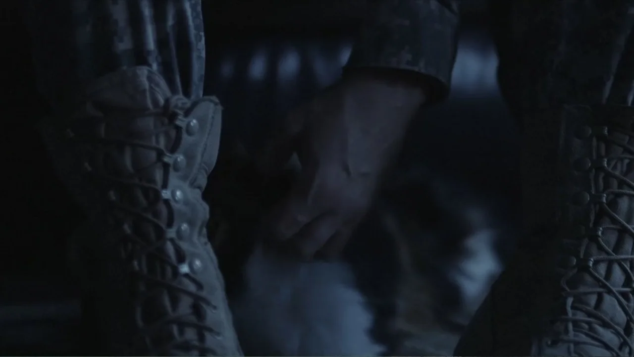 Death of a Child (2017) movie still: closeup — A dark interior space with a person wearing tactical camouflage pants and lace-up boots. One hand is…