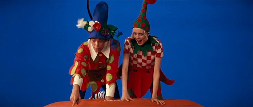The Boy Friend (1971) movie still: medium — Two individuals dressed in colorful elfin costumes stand before a bright blue studio backdrop. They are…