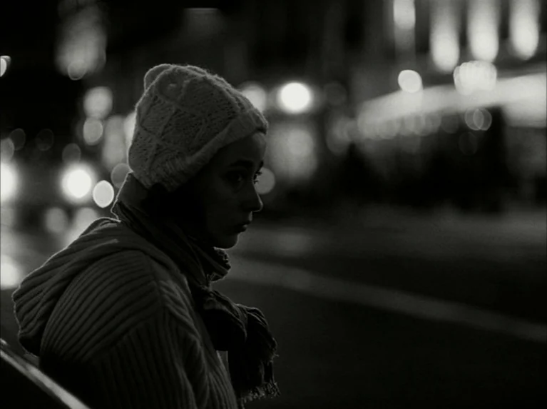 Eloge de L’amour (2001) movie still: medium — An urban sidewalk at night with blurry distant city lights forming a dark backdrop. A young woman in a…