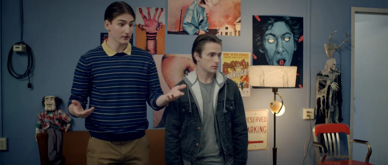 Elevator Game (2023) movie still: medium — A room with blue walls features several graphic posters and a floor lamp. Two young men stand in front of…