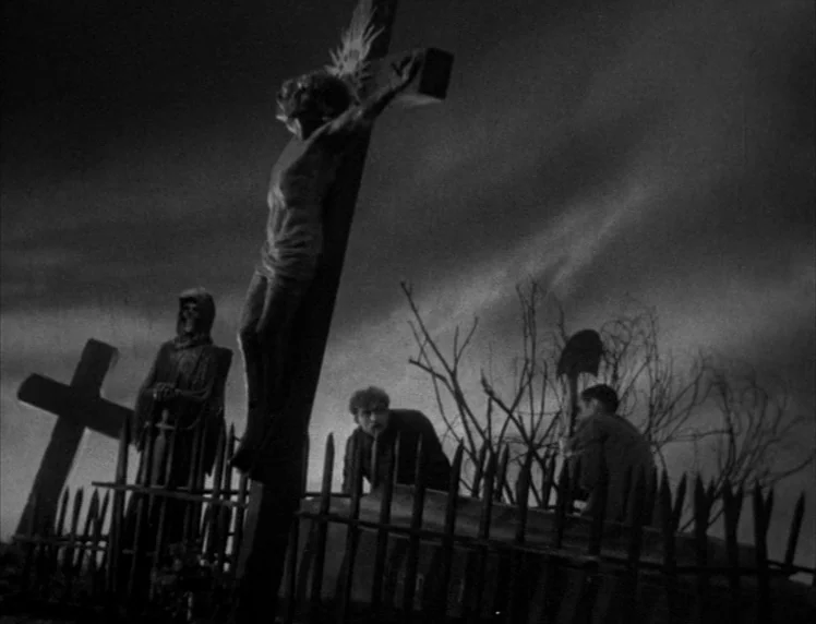 Frankenstein movie still: wide — A night cemetery features a prominent wooden crucifix and a skeletal grave marker statue. Two men crouch near a…