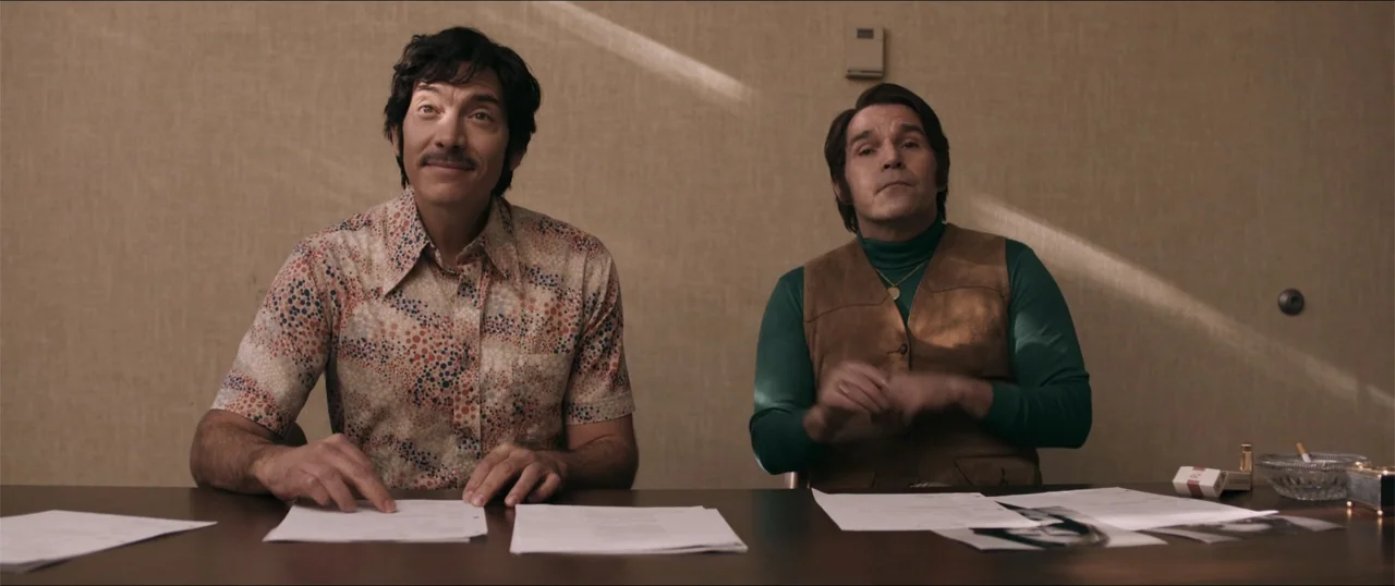 Woman of the Hour (2024) movie still: medium — An interior office with textured beige walls and a dark table. Two men in 1970s attire sit at the…