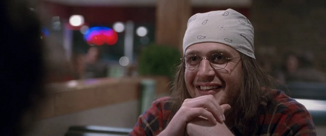 The End of the Tour (2015) movie still: closeup — A diner booth with dark upholstery and wood trim at night. A young man with a bandana and glasses…