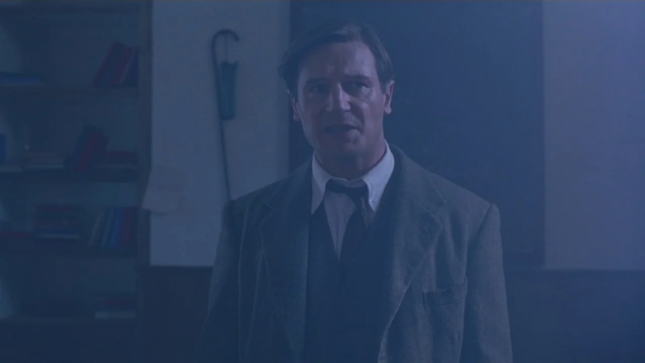 Michael Collins (1996) movie still: medium — A dark study contains a bookshelf filled with books and a wooden wall paneling. A middle-aged man in a…