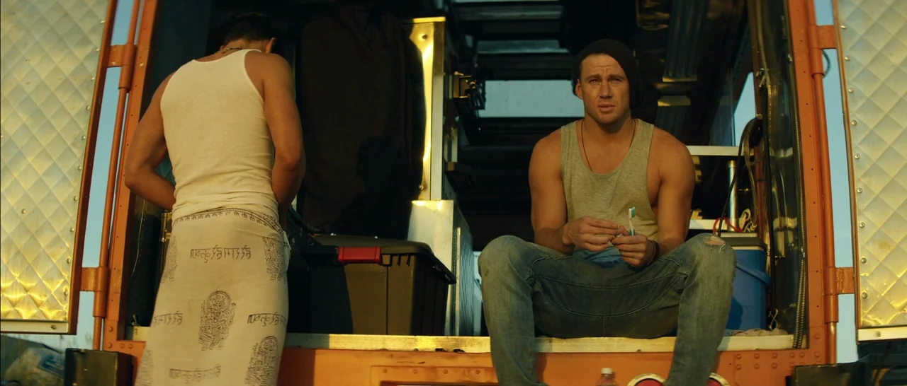 Magic Mike XXL (2015) movie still: medium — The open rear of a large metal trailer serves as a makeshift living area. A man in a beanie sits on the…