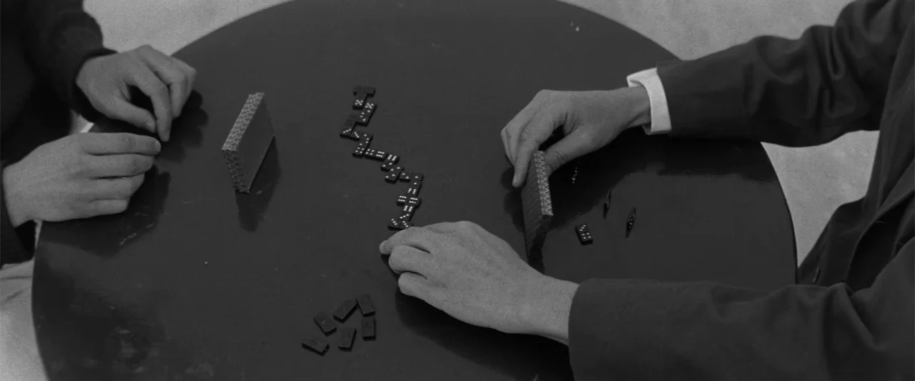 Jules and Jim (1962) movie still: medium — A top-down view of a round dark table with dominoes laid out. Two individuals are placing and moving…