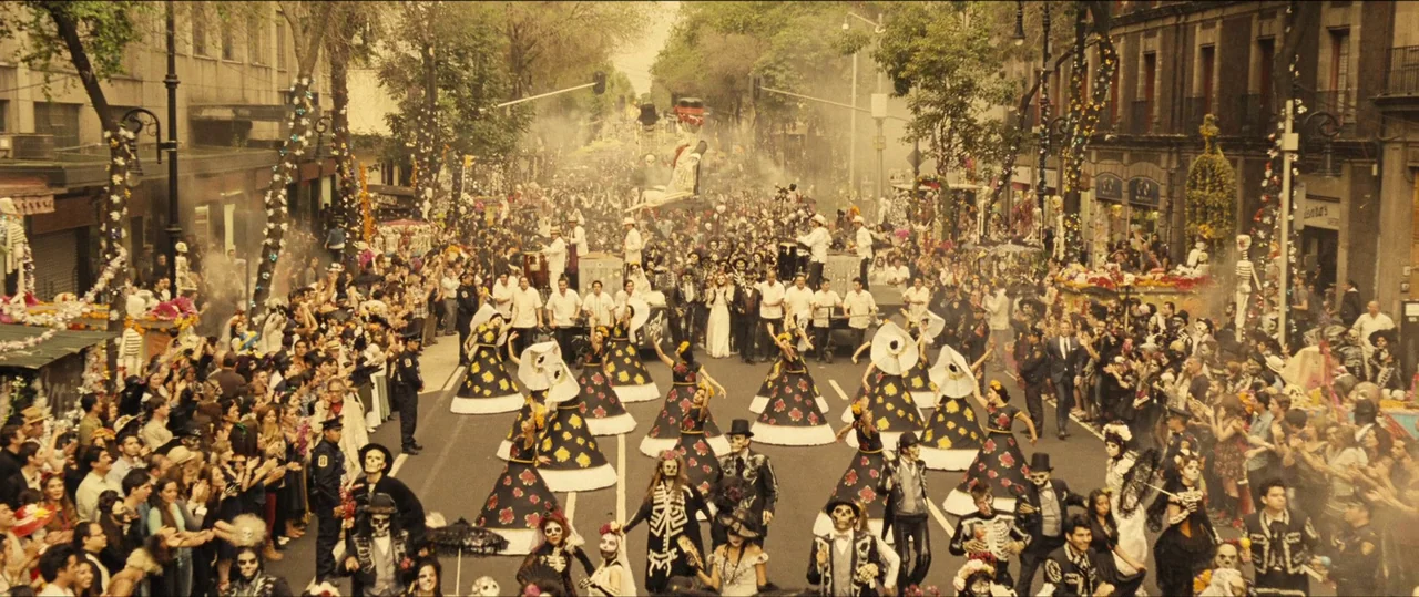 Spectre (2015) movie still: wide — A crowded metropolitan street features a large public parade with dancers in elaborate costumes. People line the…