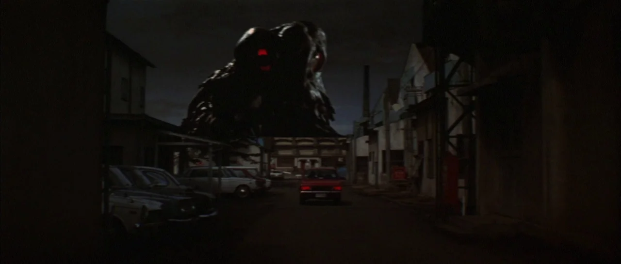 Godzilla Vs Hedorah (1971) movie still: wide — A dark, narrow alleyway at night contains several parked cars and low-rise industrial buildings. A…