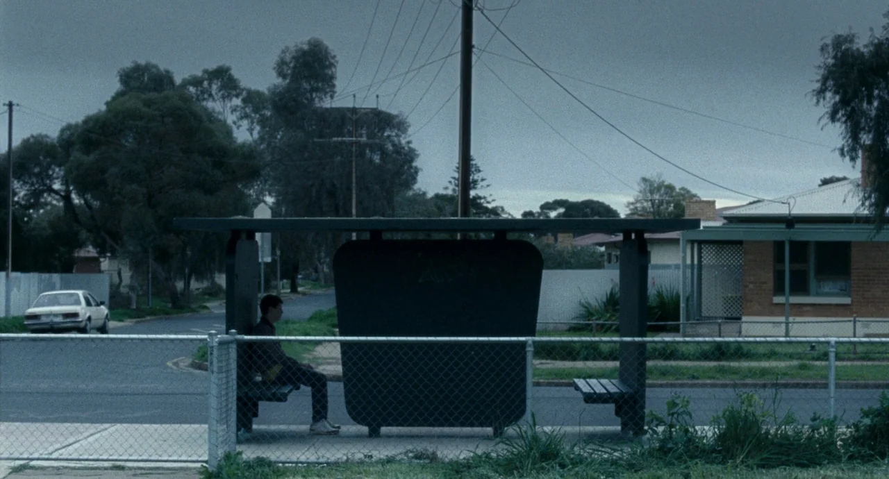 Snowtown (2011) movie still: wide — A suburban bus stop sits beside a quiet street with a residential house behind. A solitary male teenager sits on…