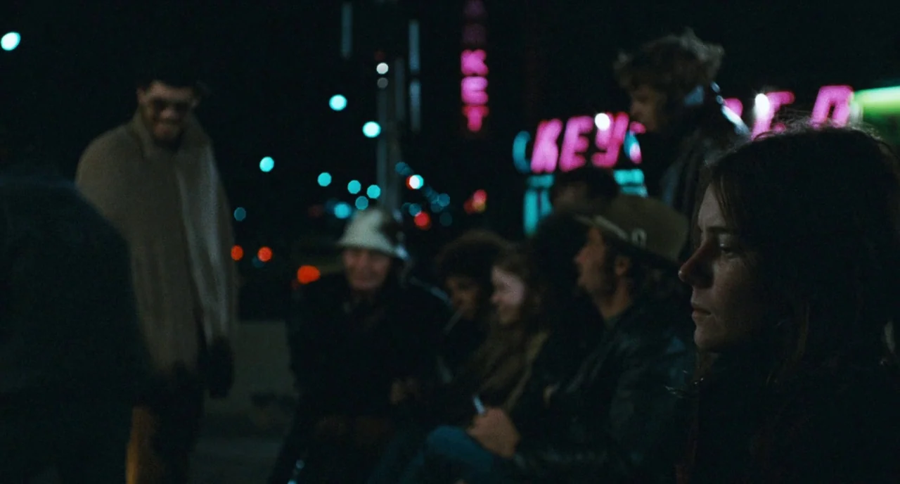 The Panic in Needle Park (1971) movie still: medium — A group of people gathers in a dark asphalt lot at night under neon. A woman stands in the…